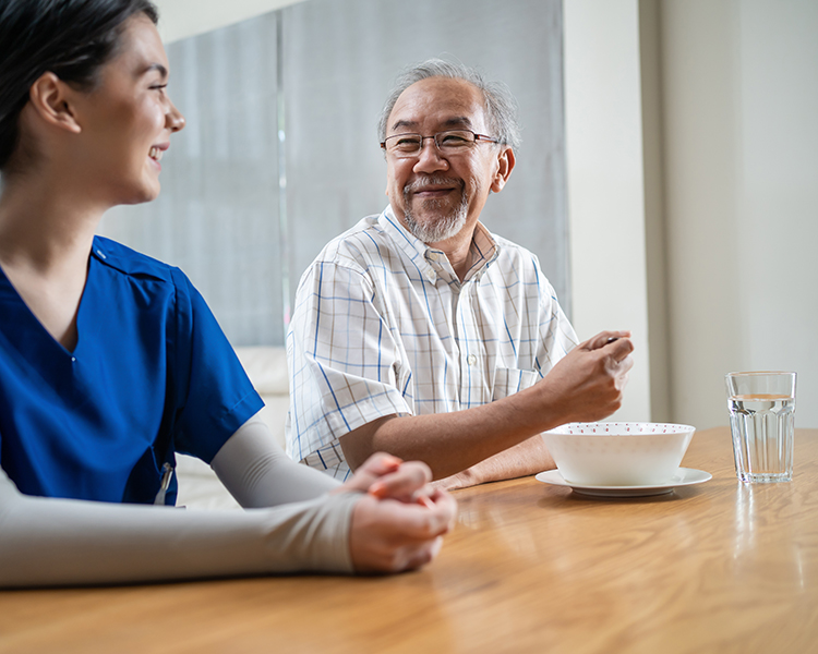 A caregiver in blue scrubs and an elderly man share a joyful conversation at a table, with the elderly man holding a spoon and a bowl in front of him, highlighting companionship and care during mealtime.