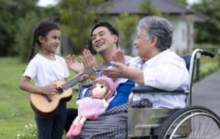 Family spending time together outdoors as a caregiver supports an elderly woman in a wheelchair while a young girl plays ukulele, highlighting holiday caregiving balance.